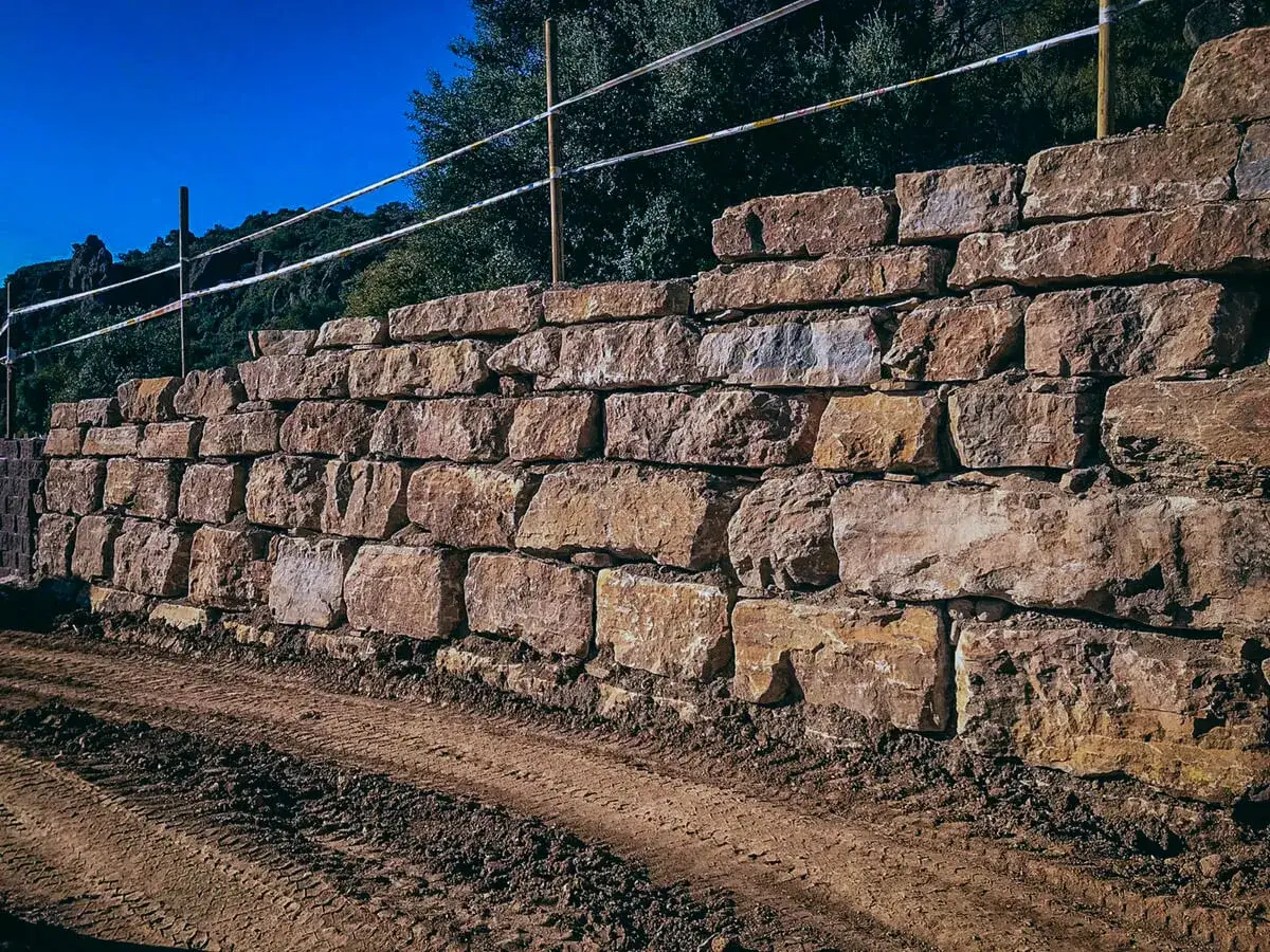 Un muro escollera de grandes rocas de tonos ocres y rojizos, con tierra removida y huellas de tractor en primer plano.