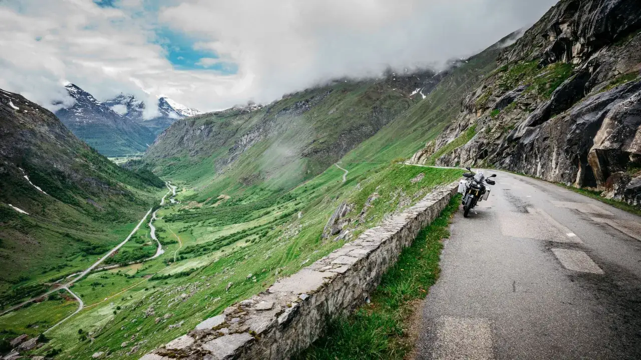 Moto parcheggiata su una strada alpina tortuosa, con una valle verdeggiante e montagne innevate sullo sfondo. Un viaggio verso il passo più alto d'Europa.