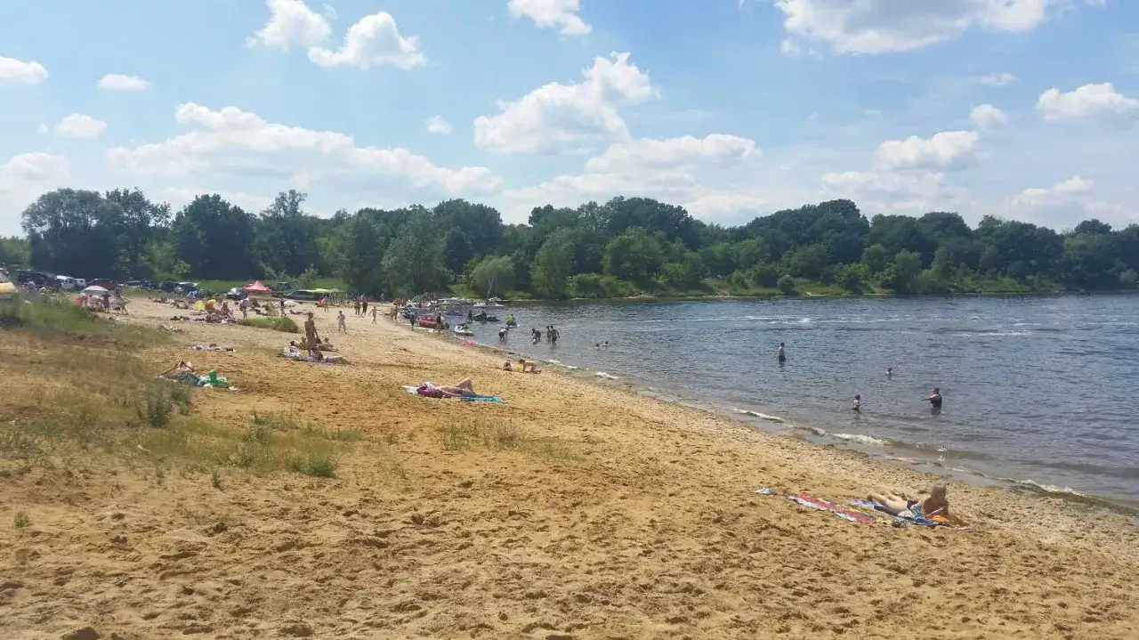 Słoneczny dzień nad **rzeczyce jezioro**. Ludzie odpoczywają na piaszczystej plaży, kąpią się i pływają.