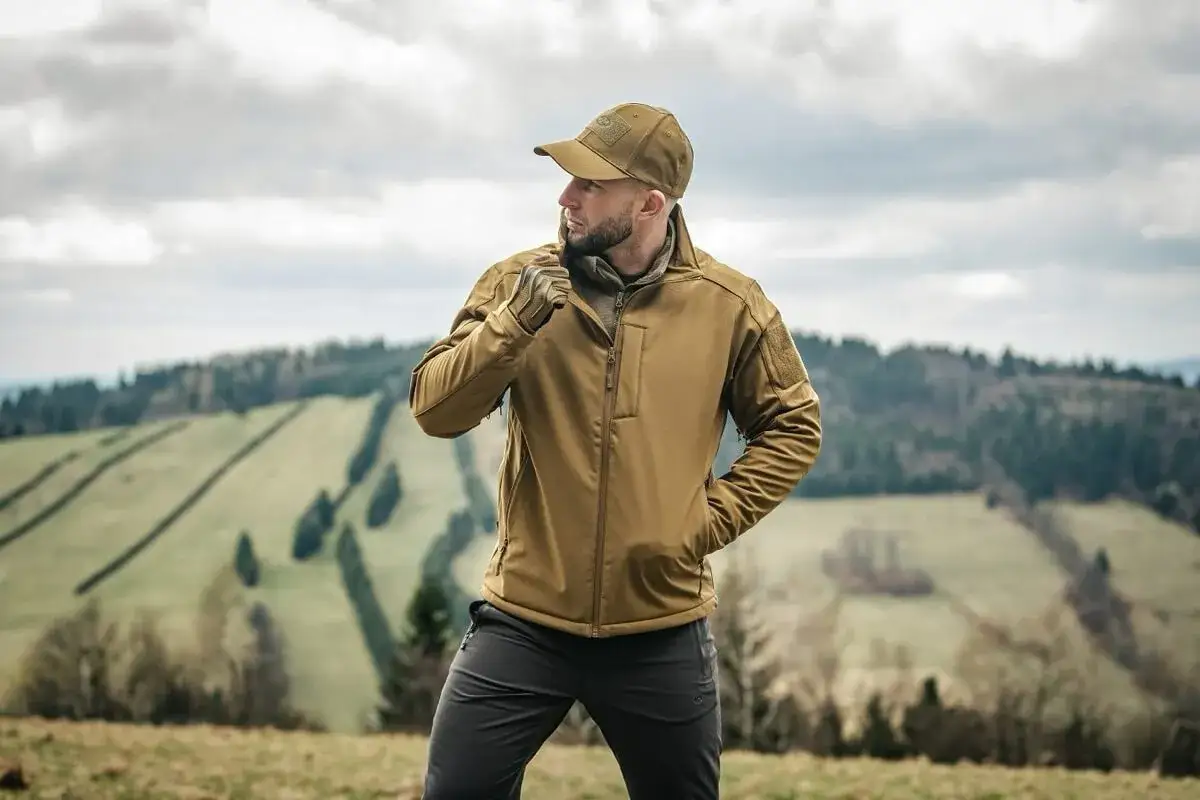 Mężczyzna w beżowej kurtce softshell co to jest, pozuje na tle górskiego krajobrazu.