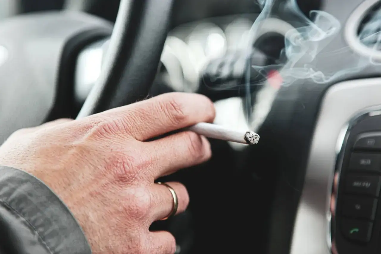 Mano con cigarrillo encendido y humo saliendo, mientras se puede fumar conduciendo.