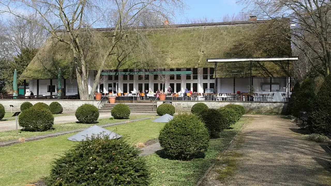 Das Teehaus im Englischen Garten Berlin mit seinem Reetdach und der Terrasse, auf der G&auml;ste sitzen, ist von gepflegten Rasenfl&auml;chen und B&uuml;schen umgeben.