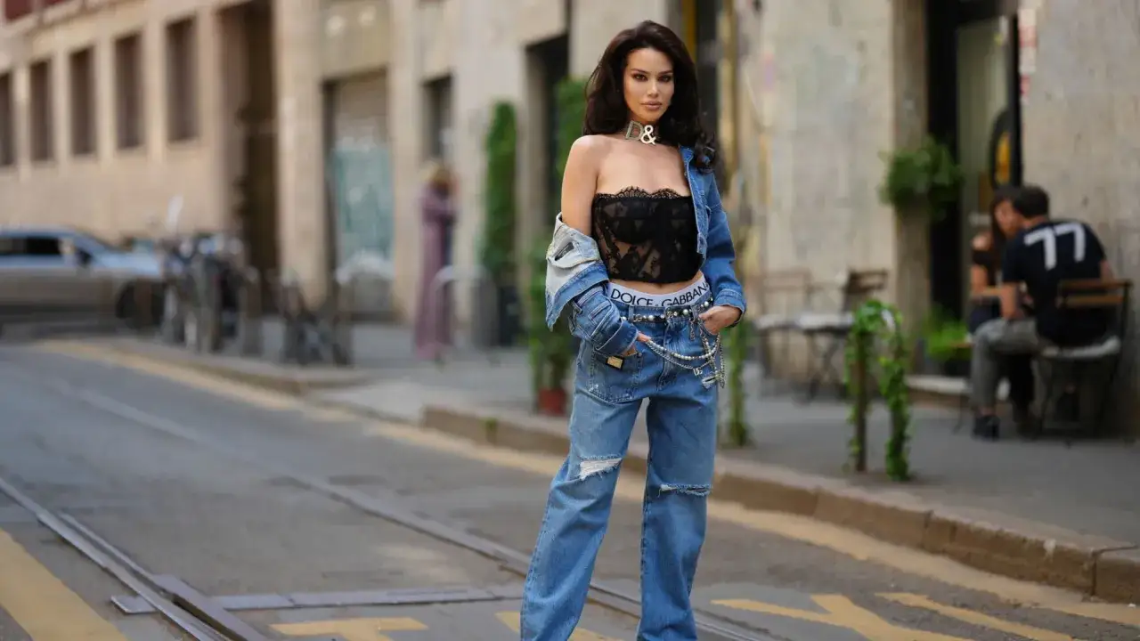 Donna con jeans strappati e giacca di jeans aperta, mostrando un reggiseno a vista in pizzo nero e slip con logo.