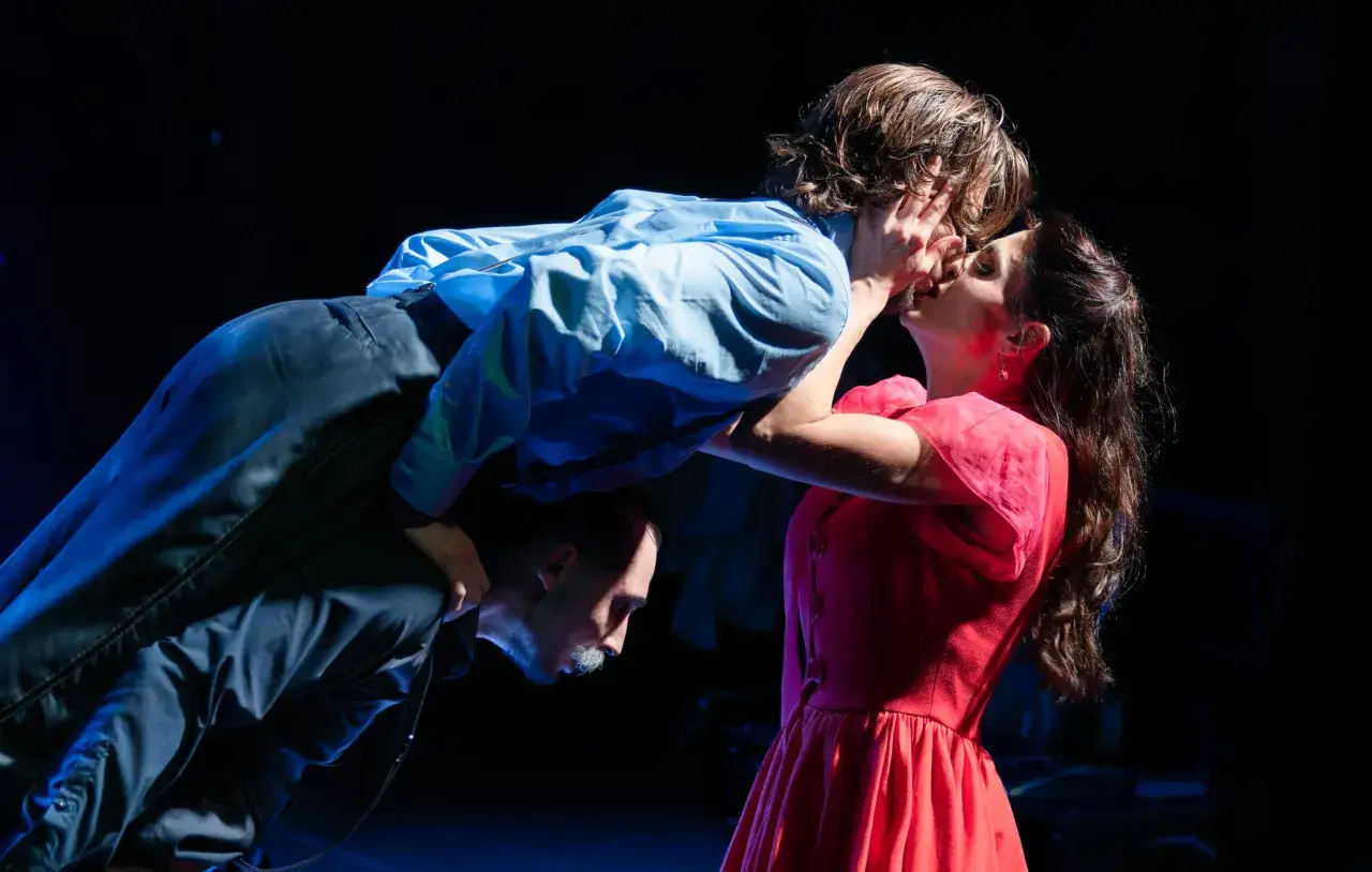 Intensywna scena z teatru. Mężczyzna w niebieskiej koszuli obejmuje kobietę w czerwonej sukience, która go całuje. Pod nimi klęczy inny mężczyzna.