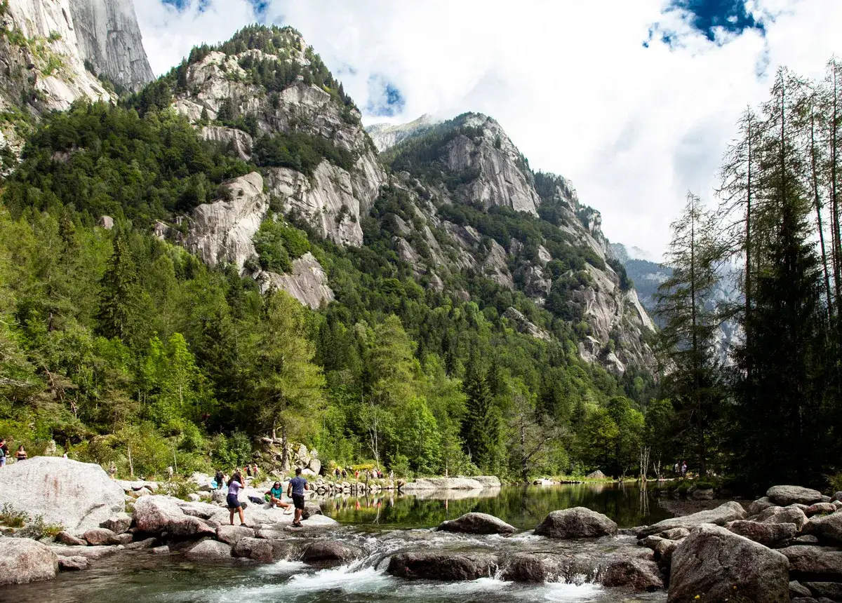 Gente in escursione nel **trekking Val di Mello**, tra rocce, boschi e un lago alpino.