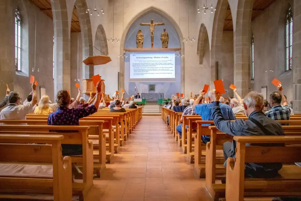 Menschen in einer Kirche stimmen mit roten Karten ab. Ein Bildschirm zeigt Text &uuml;ber die katholische Soziallehre.