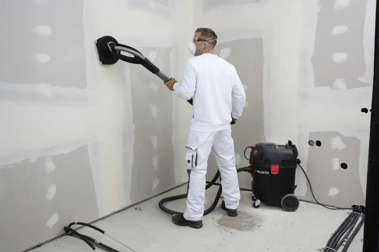 Un operaio con maschera e occhiali sta carteggiando un muro per imbiancare, collegato a un aspiratore W&uuml;rth per raccogliere la polvere.