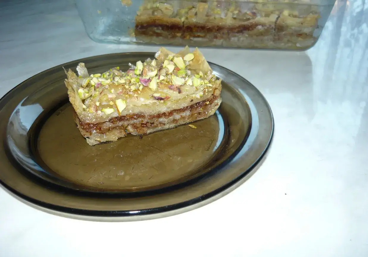Słodka, warstwowa baklawa turecka z pistacjami, podana na ciemnym talerzu. W tle widać resztę deseru w szklanym naczyniu.