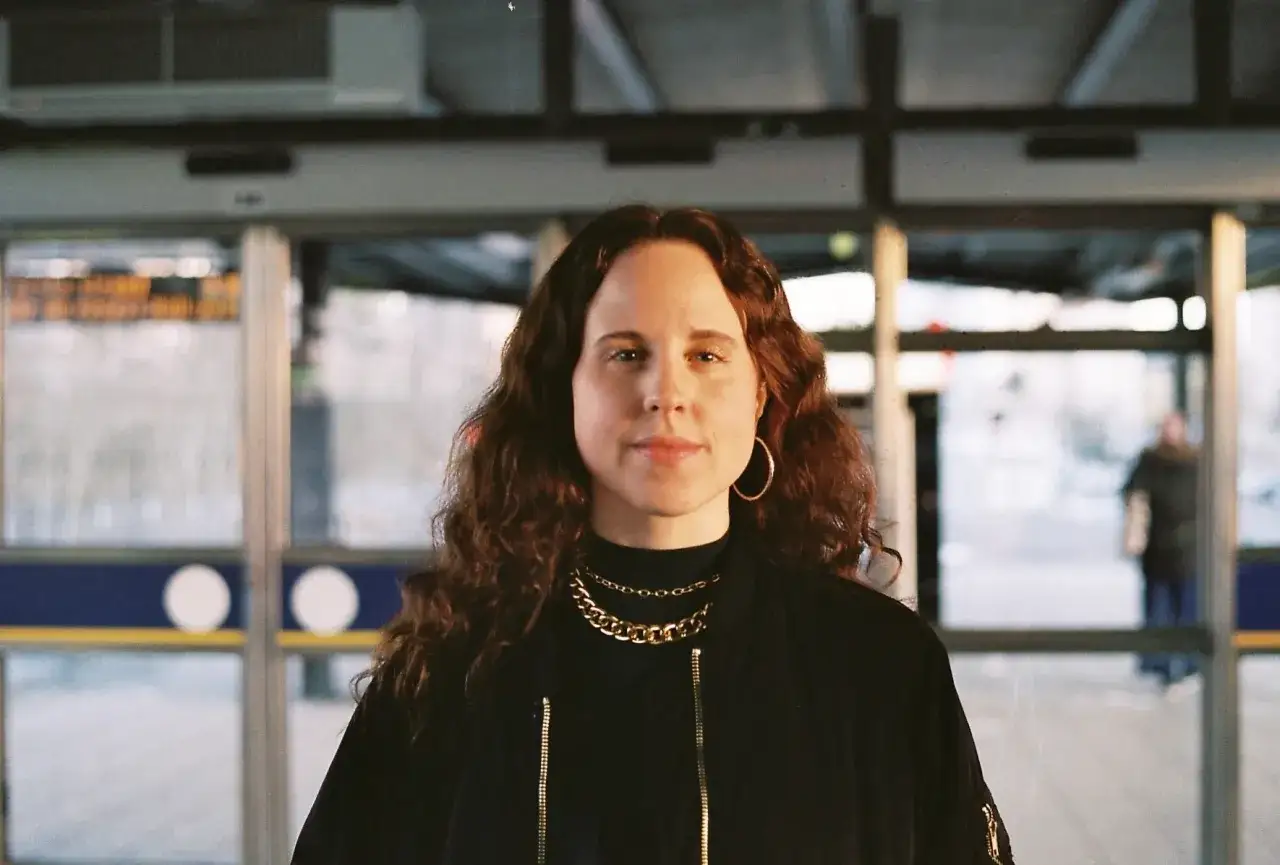 Dina Ekborg med lockigt h&aring;r och guldkedjor, st&aring;r framf&ouml;r glasd&ouml;rrar.