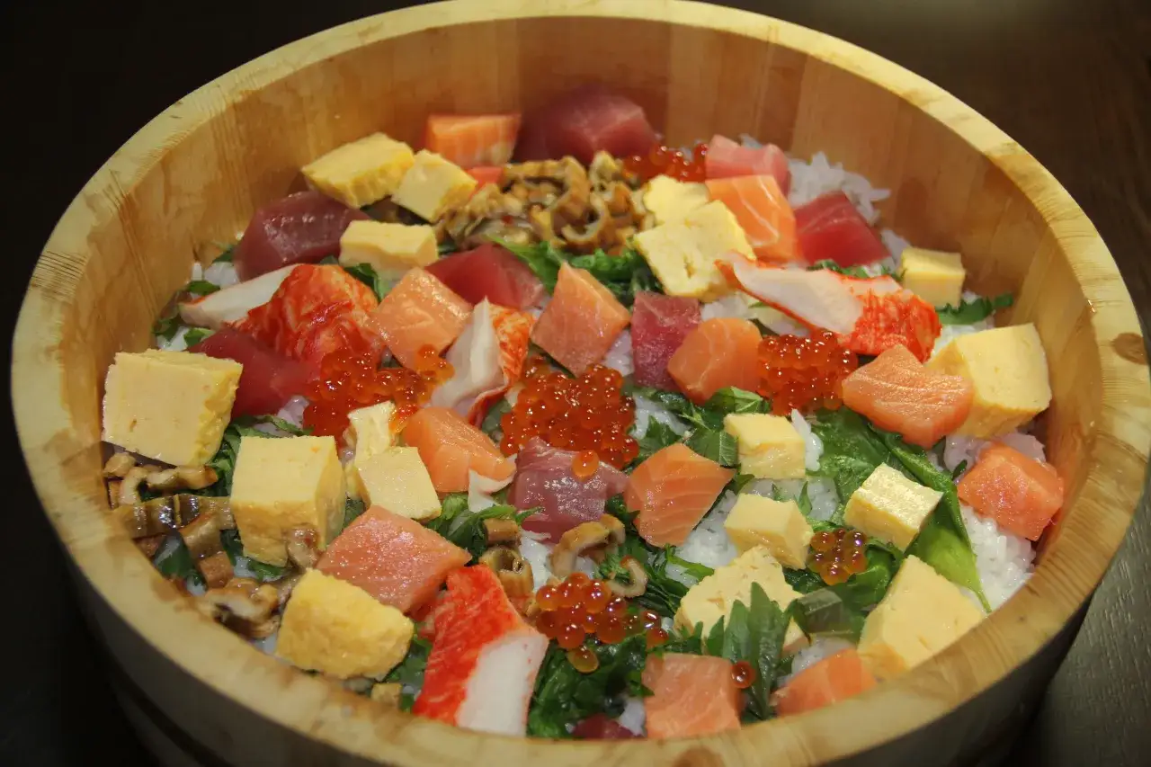 Kolorowe chirashi zushi w drewnianej misie: kawałki łososia, tuńczyka, omletu tamago, ikry i surimi na ryżu.