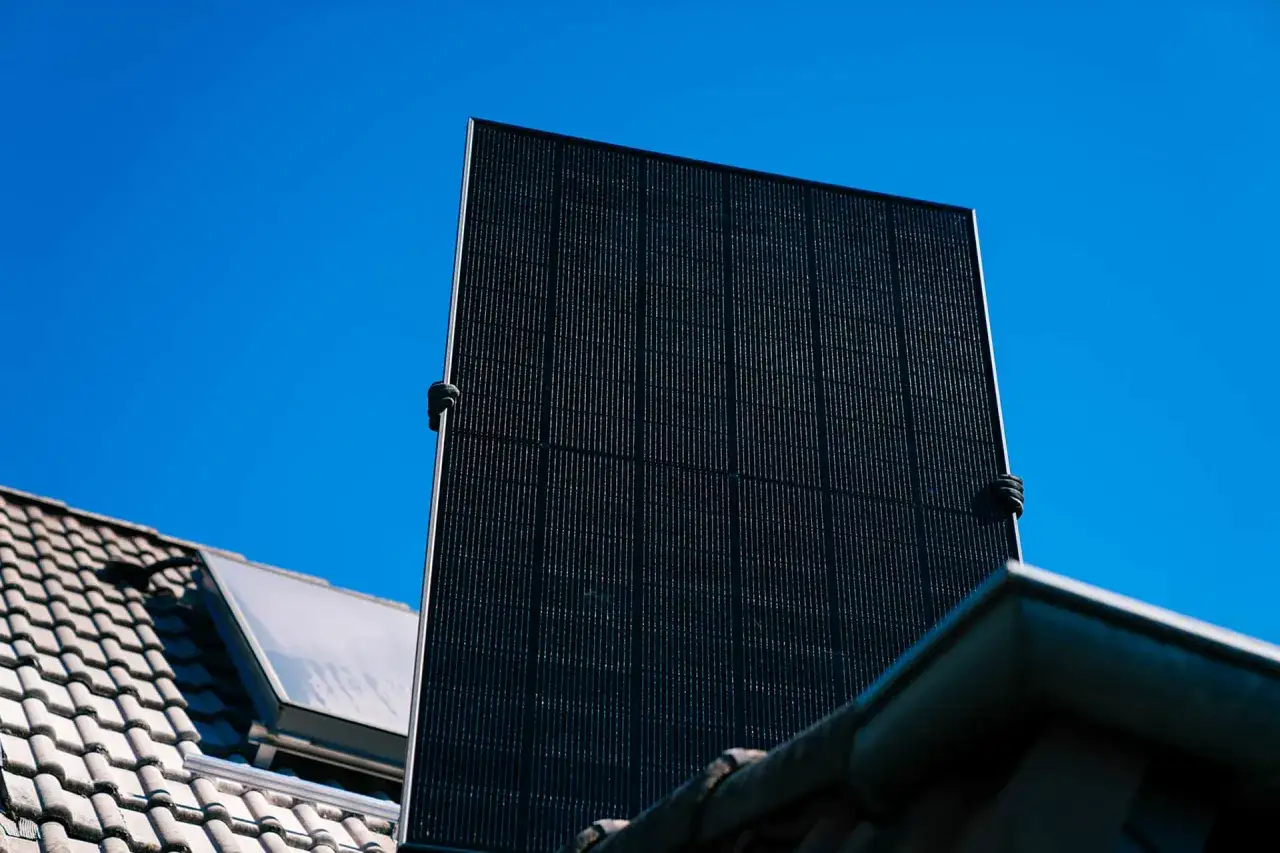 Tv&aring; h&auml;nder h&aring;ller upp en stor, m&ouml;rk solpanel mot en klarbl&aring; himmel. Bredvid syns ett tegeltak och en solcell.
