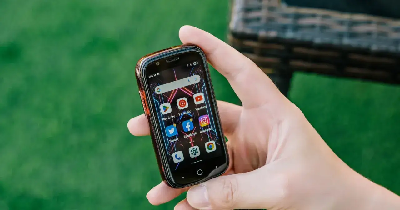 Dłoń trzyma najmniejszy smartfon z ikonami aplikacji, w tym YouTube i Facebook, na tle zielonej trawy.