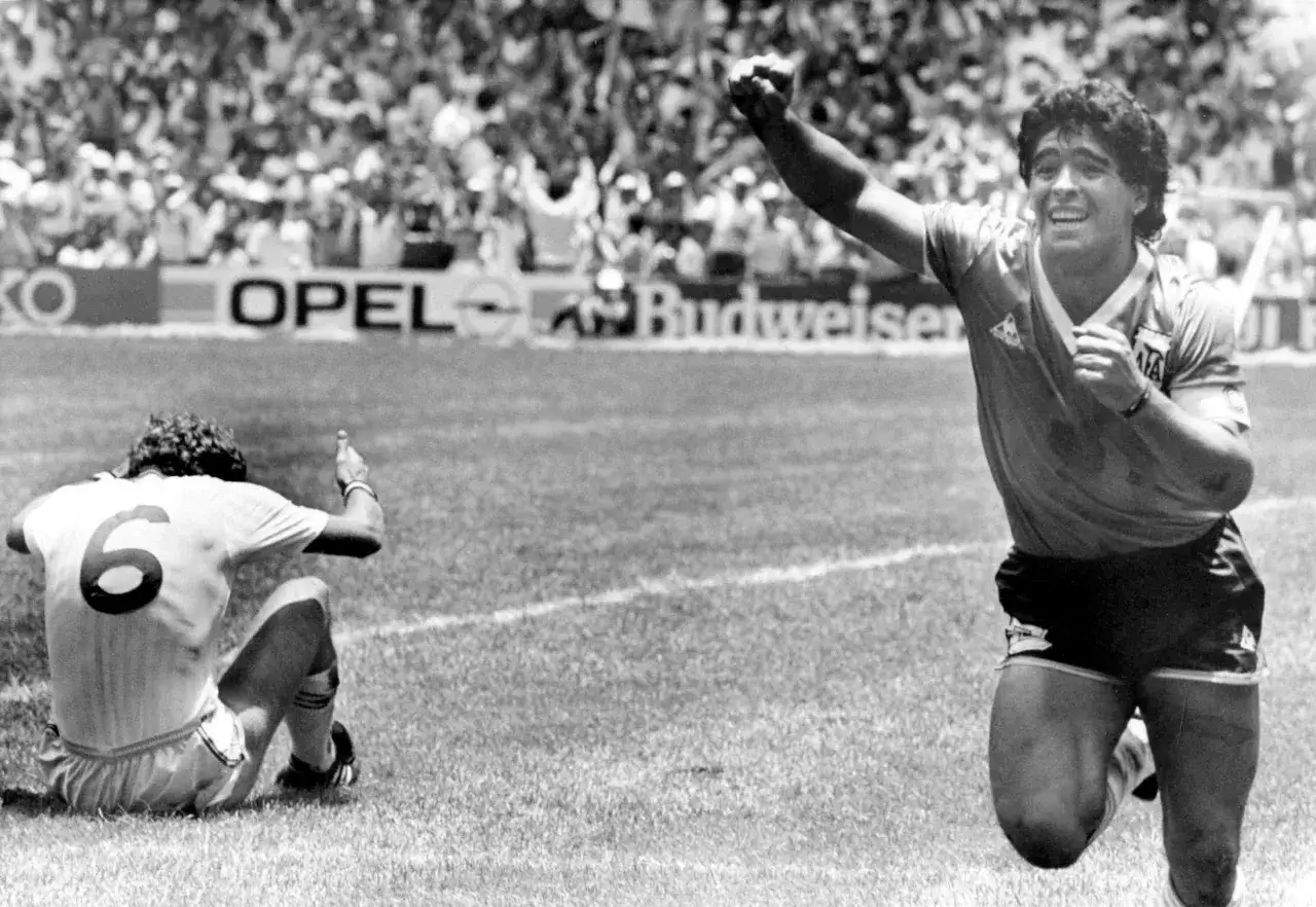Diego Maradona świętuje bramkę, podczas gdy przeciwnik z numerem 6 siedzi na boisku. Dwóch znanych piłkarzy w akcji.