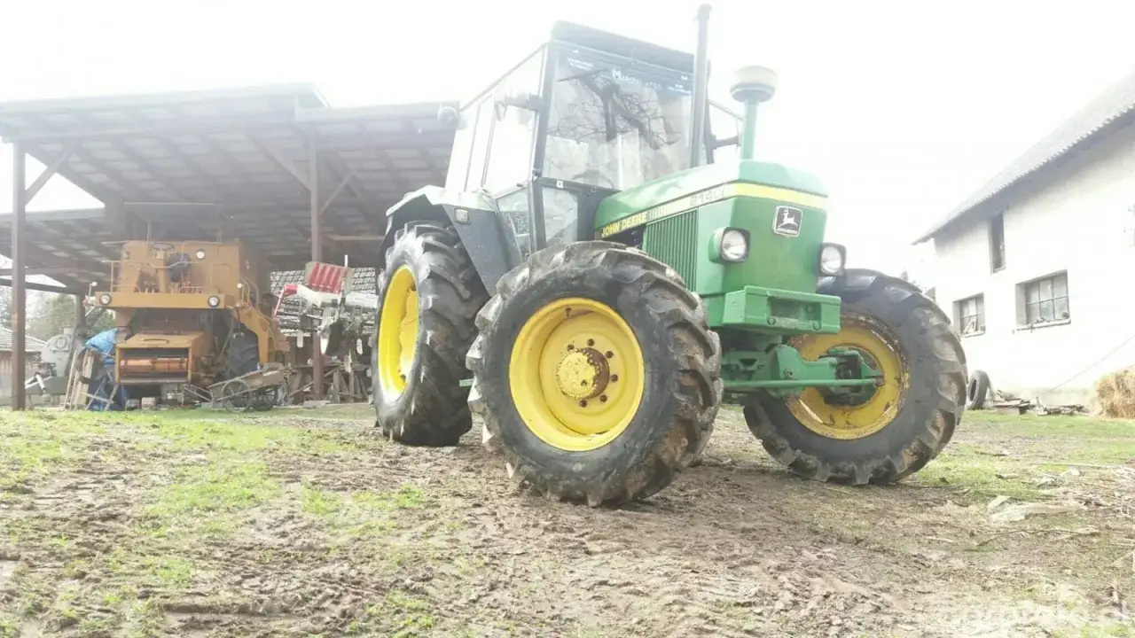 Zielony John Deere 2140 z żółtymi felgami stoi na błotnistym podwórku obok stodoły i żółtej maszyny rolniczej.