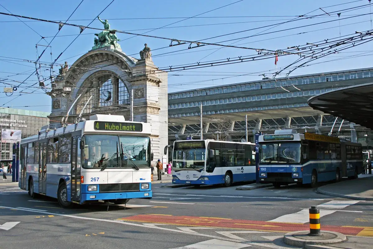 Trolleybuses wait for passengers at the historic Lucerne train station, with its grand archway and clock tower.