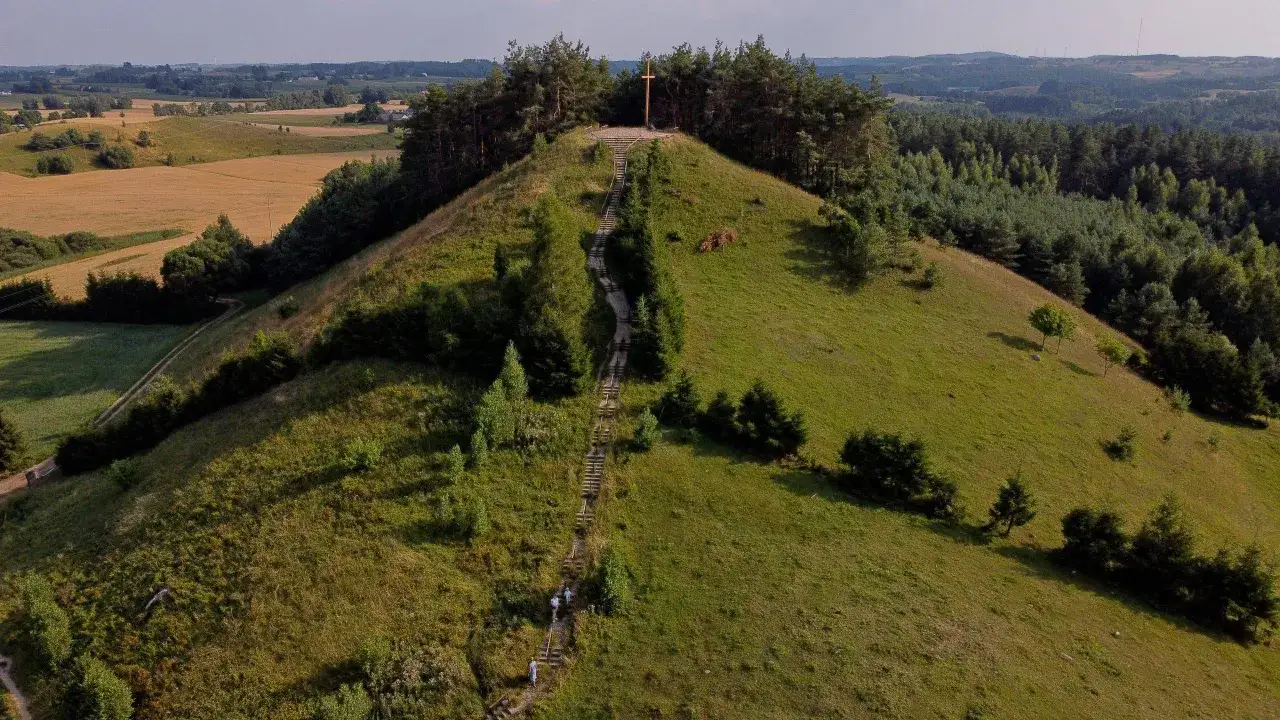 Wzgórze porośnięte trawą i drzewami, z drewnianym krzyżem na szczycie. Długa ścieżka ze schodami prowadzi na górę. Cisowa góra w letnim krajobrazie.