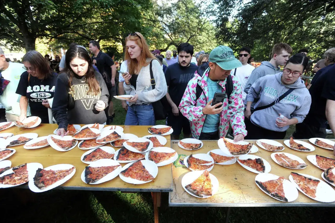 Ludzie wybierają kawałki pizzy z ogromnego stołu. Widać wiele talerzy z pizzą, jakby była to pizza 100cm.