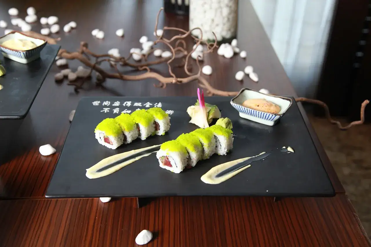 Jak jeść sushi? Na czarnej desce ułożono rolki sushi z zielonym ikrą, sos, wasabi i marynowany imbir.