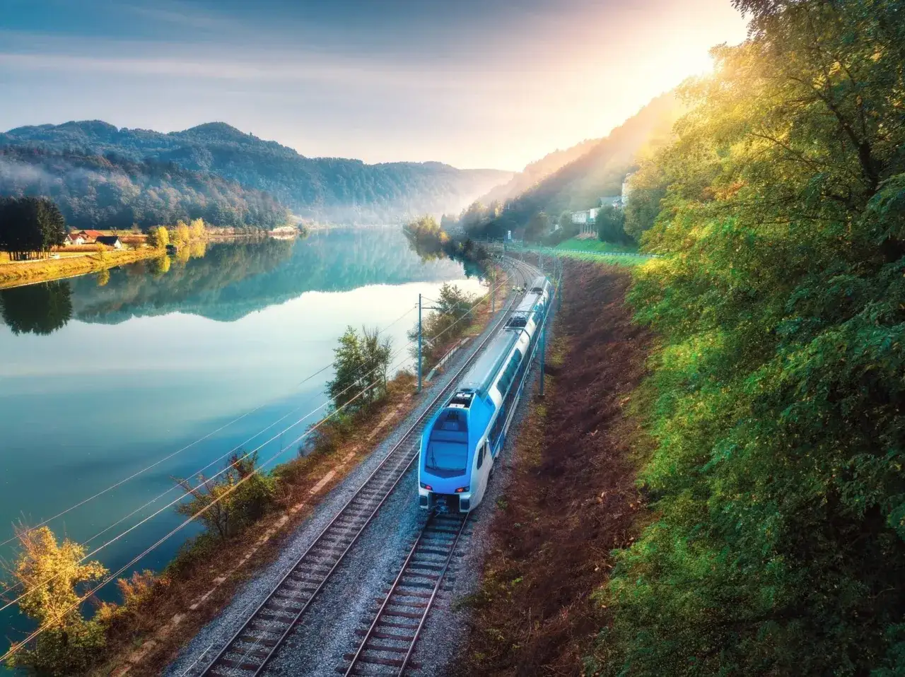 Ein moderner Zug fährt auf dem Bahnnetz Frankreich entlang eines ruhigen Flusses, umgeben von grünen Hügeln und im warmen Licht der Morgensonne.