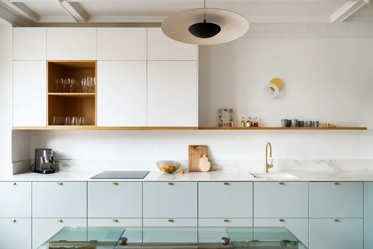 Une cuisine moderne aux fa&ccedil;ades bleu pastel et aux accents de bois. Des verres sont rang&eacute;s dans une niche en bois, tandis qu'un plan de travail en marbre accueille une cafeti&egrave;re et un &eacute;vier dor&eacute;.