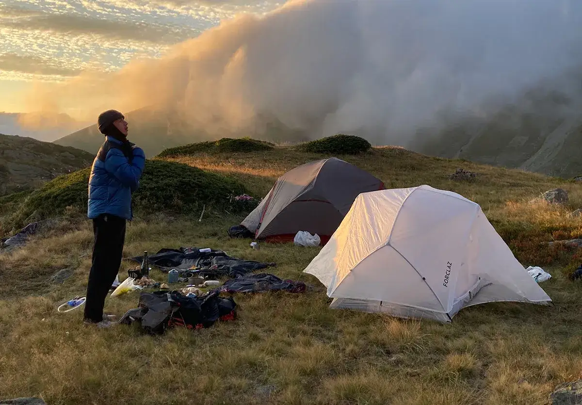 Une personne se tient pr&egrave;s d'une aire de bivouac avec deux tentes, des sacs et du mat&eacute;riel de camping, au lever du soleil dans les montagnes.