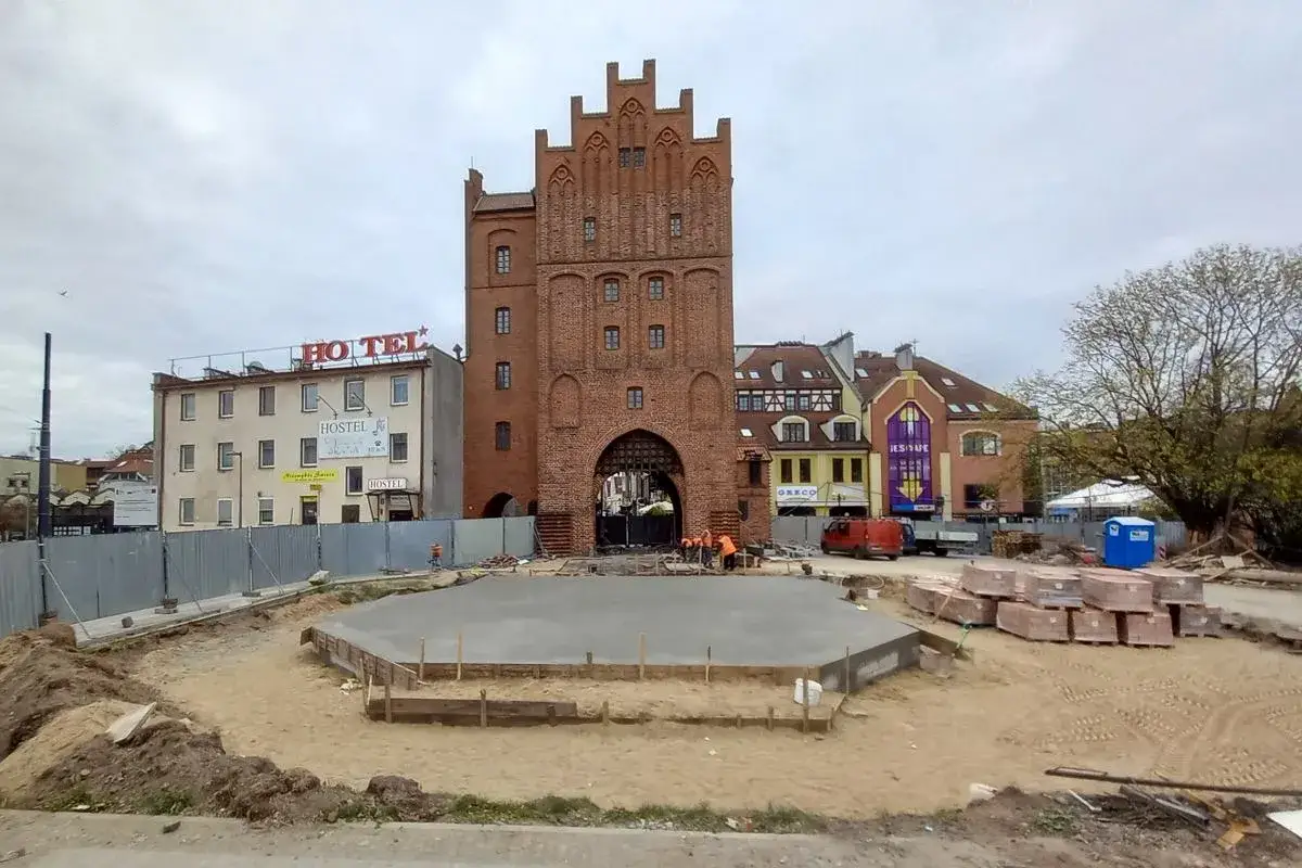 Plac budowy w polskim Carcassonne. W tle ceglana wieża, hotel i kolorowe kamienice. Trwają prace przy wylewce betonu.