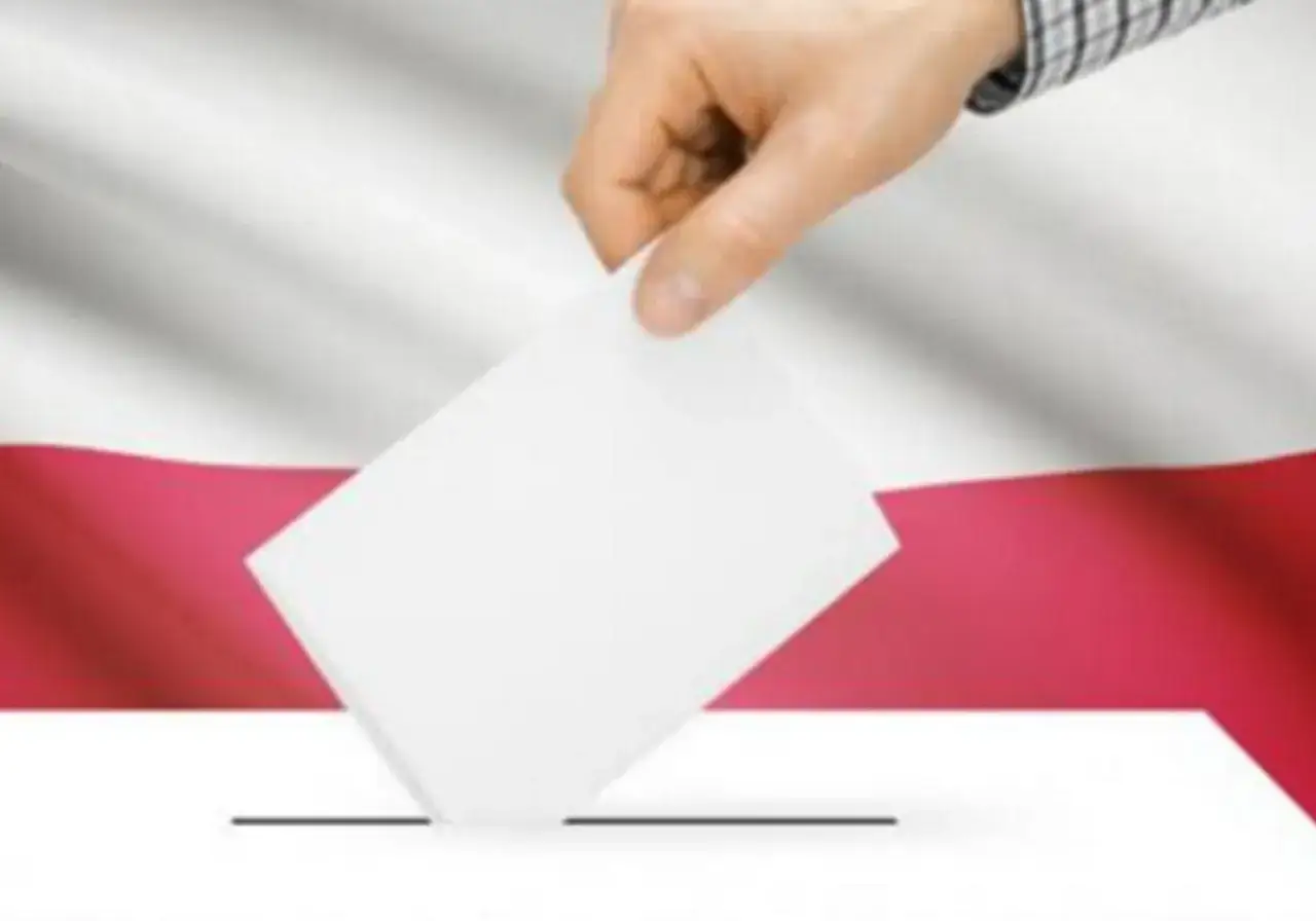 Dłoń wrzuca kartę do urny na tle polskiej flagi. To symbol ważnych wyborów do parlamentu.