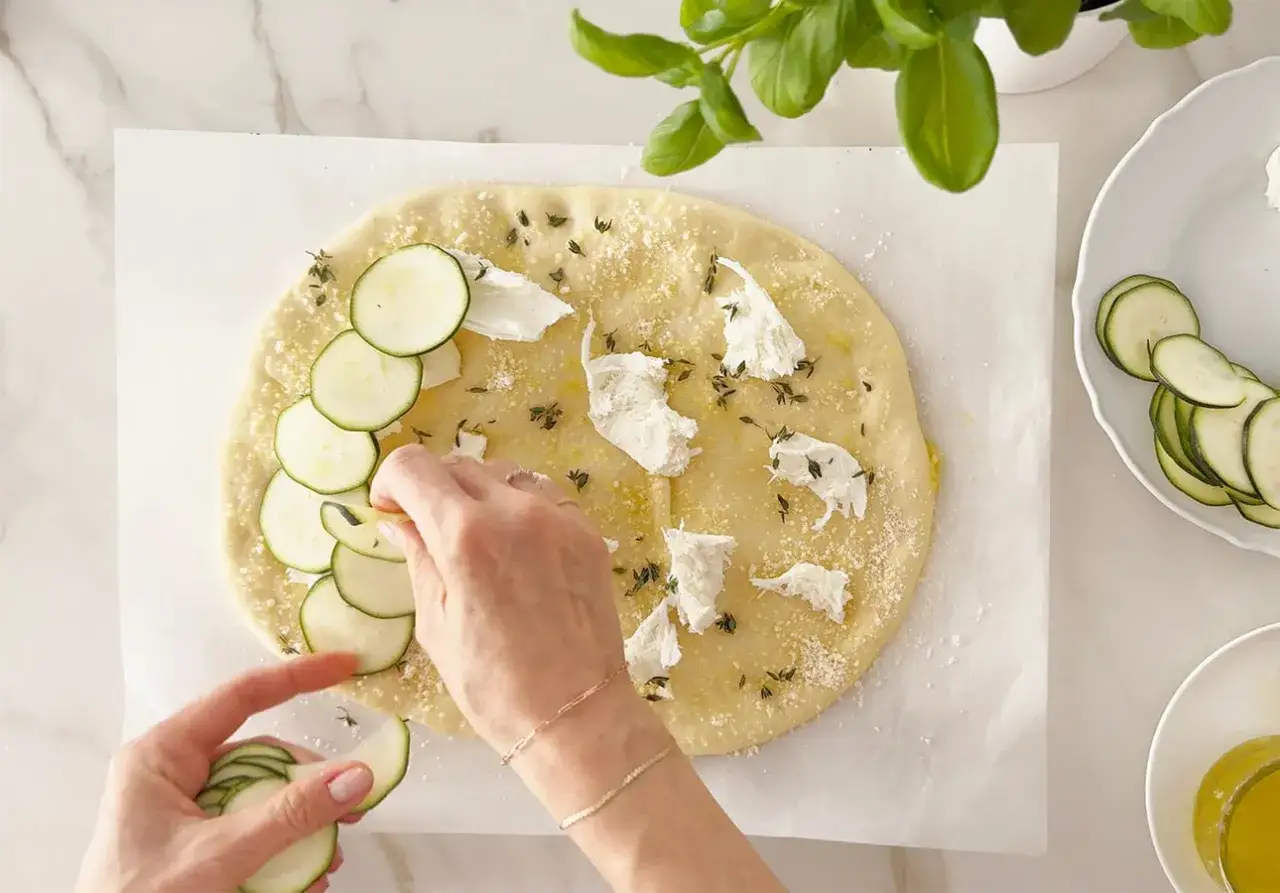 H&auml;nde belegen Pizza mit Zucchinischeiben und K&auml;se. Zucchini schmeckt auf Pizza wunderbar frisch und leicht.