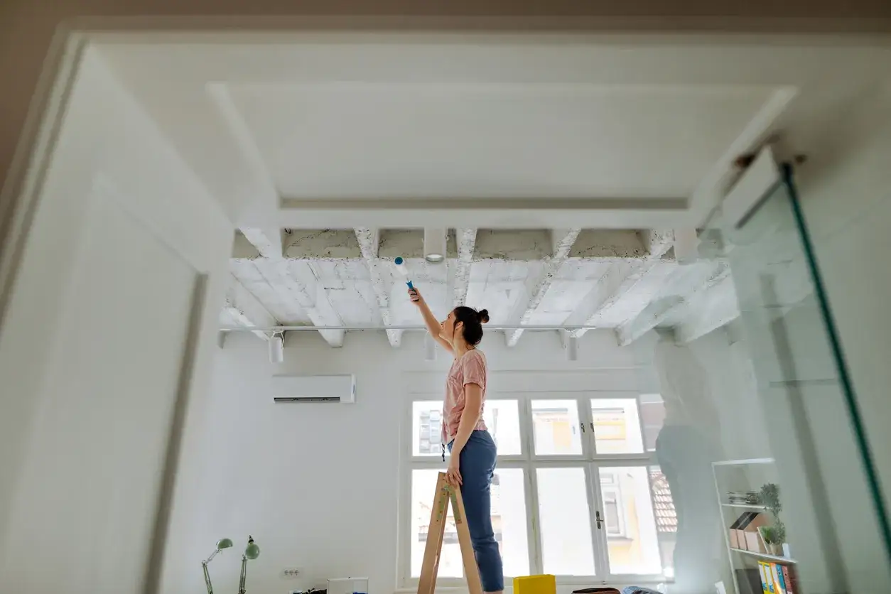 Frau streicht mit einer Rolle die Decke. Sie steht auf einer Leiter und arbeitet an den Betonbalken.
