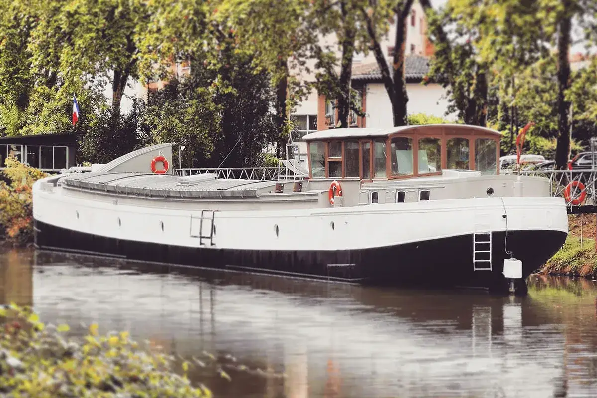P&eacute;niche blanche et noire amarr&eacute;e sur un canal bord&eacute; d'arbres. Sa longueur impressionnante sugg&egrave;re une grande capacit&eacute; de transport ou un logement spacieux.