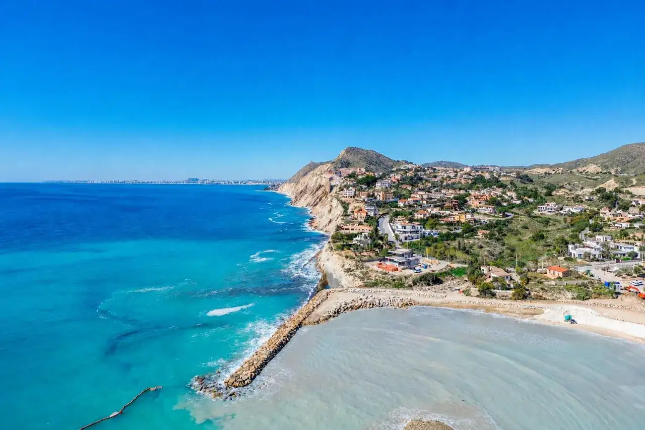 Costa azul y casas blancas en la ladera de la montaña, con una cala baeza y un rompeolas de rocas.