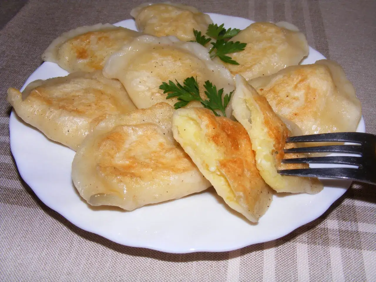 Talerz pełen pysznych, złocistych pierog&oacute;w z ziemniakami, jeden z nich jest nadziany na widelec, ozdobiony gałązką natki pietruszki.