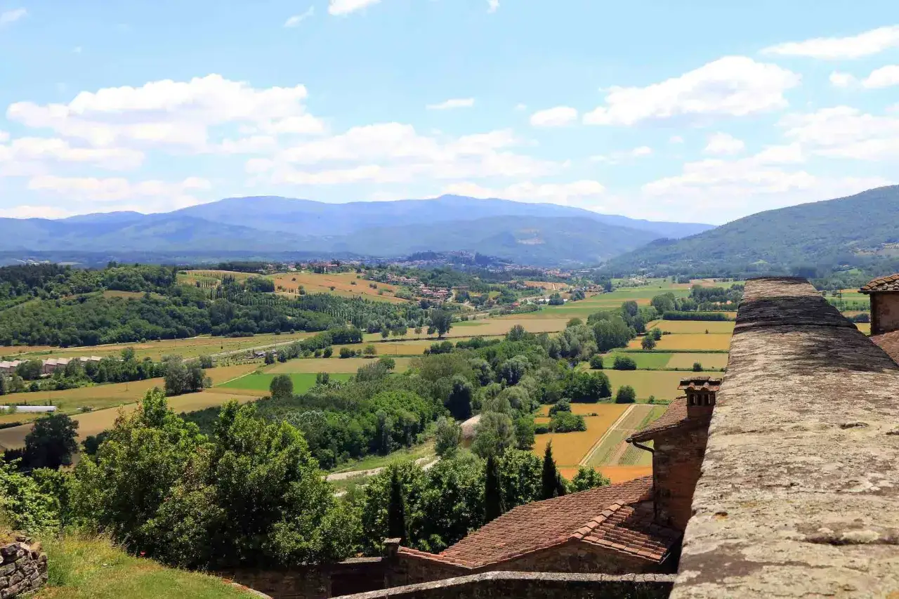 Vista panoramica di un borgo alla collina, con campi coltivati, alberi e montagne sullo sfondo.