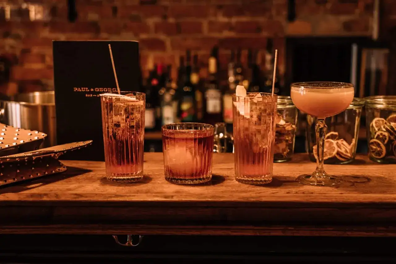 Vier Cocktails auf einer Holztheke, darunter die 10 beliebtesten Cocktails. Im Hintergrund eine Bar mit Flaschen und Gläsern.