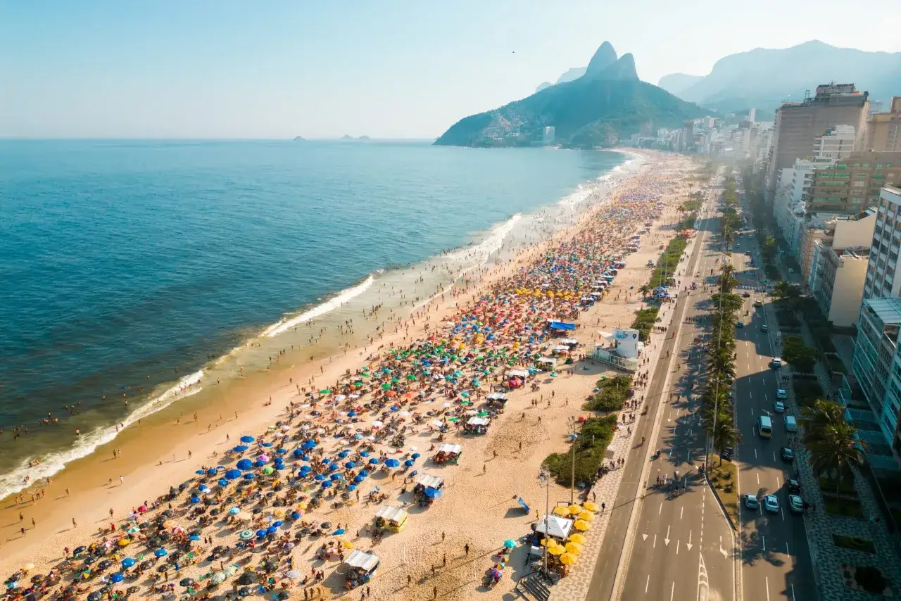 Tłum ludzi na plaży w Rio de Janeiro, z górami w tle. Czy Brazylia jest bezpieczna? Widok sugeruje spokojny dzień na plaży.