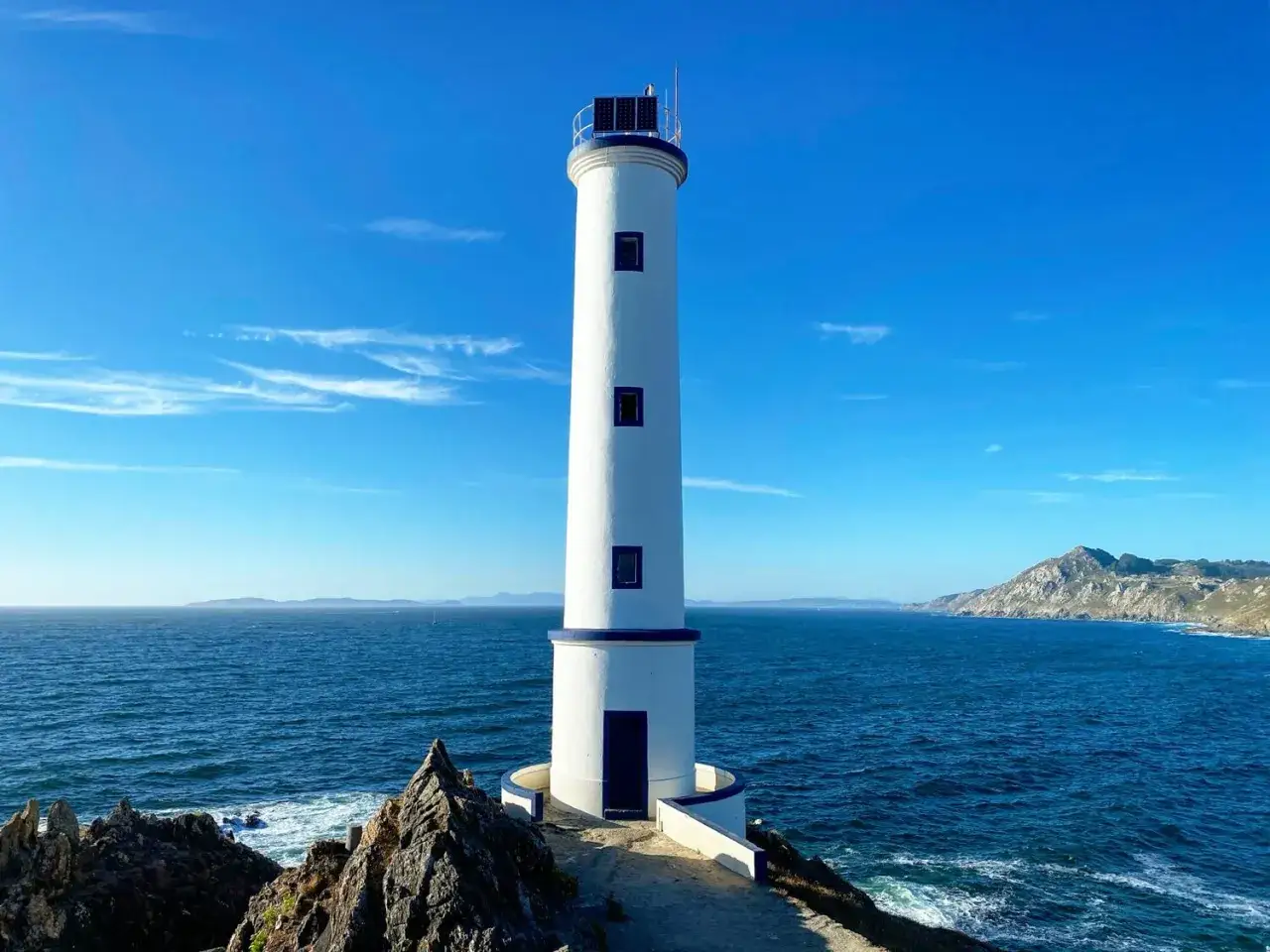 El faro de Cabo Home, blanco y azul, vigila el Atlántico bajo un cielo despejado.