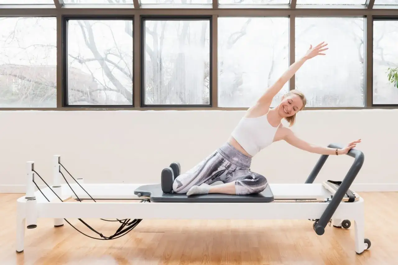 Femme souriante en plein **&eacute;tat de flow** sur un reformer Pilates, bras lev&eacute;s, &eacute;tirant son corps avec gr&acirc;ce.