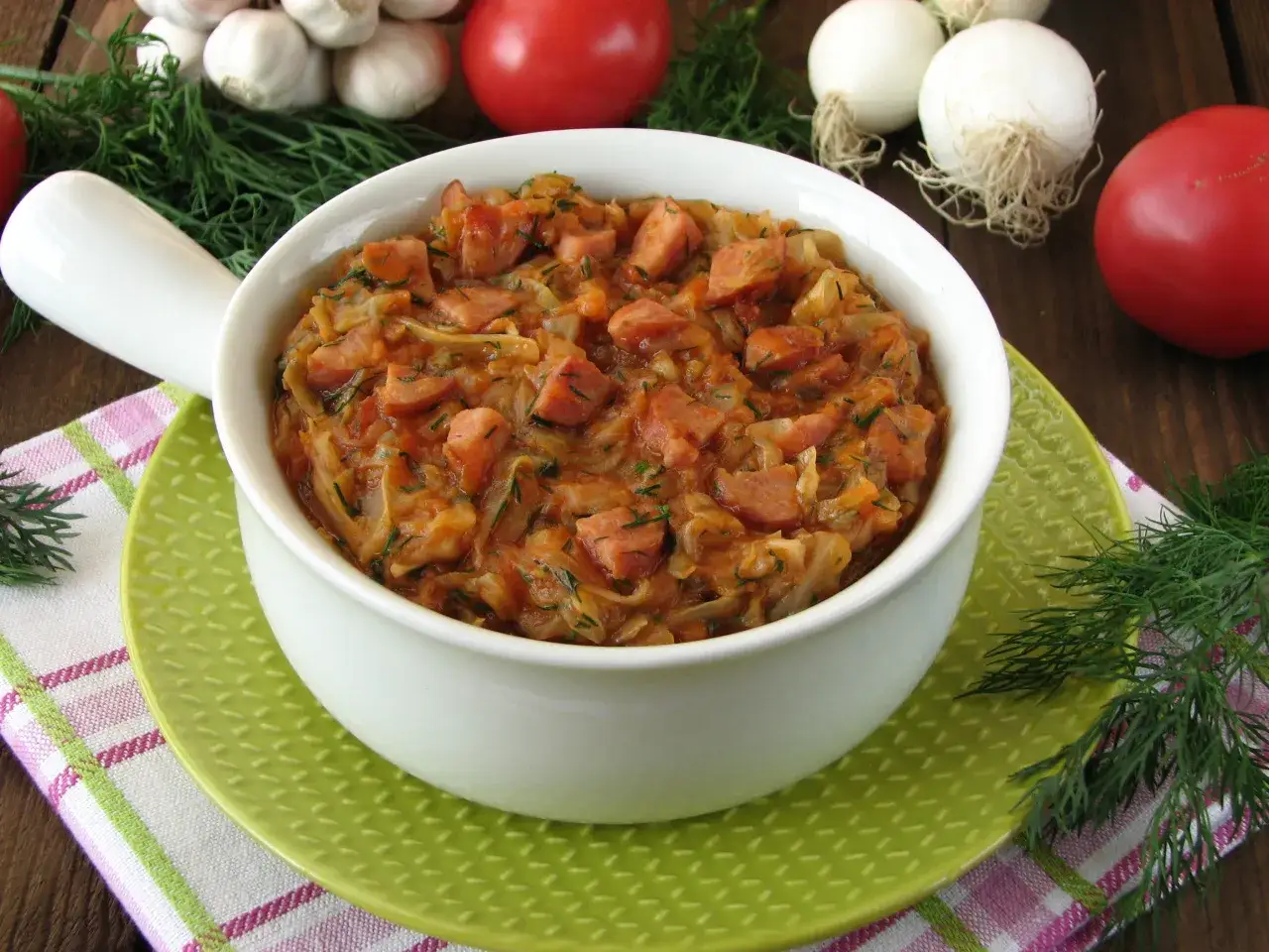 Pyszny bigos z młodej kapusty z kiełbasą i koprem, podany w białej misce na zielonym talerzyku. Obok pomidory, czosnek i cebula.