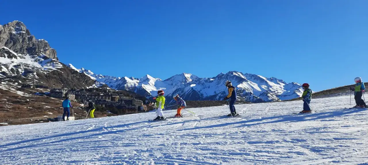 Niños aprendiendo que es el esquí alpino en una ladera nevada, con majestuosas montañas y un pueblo al fondo.