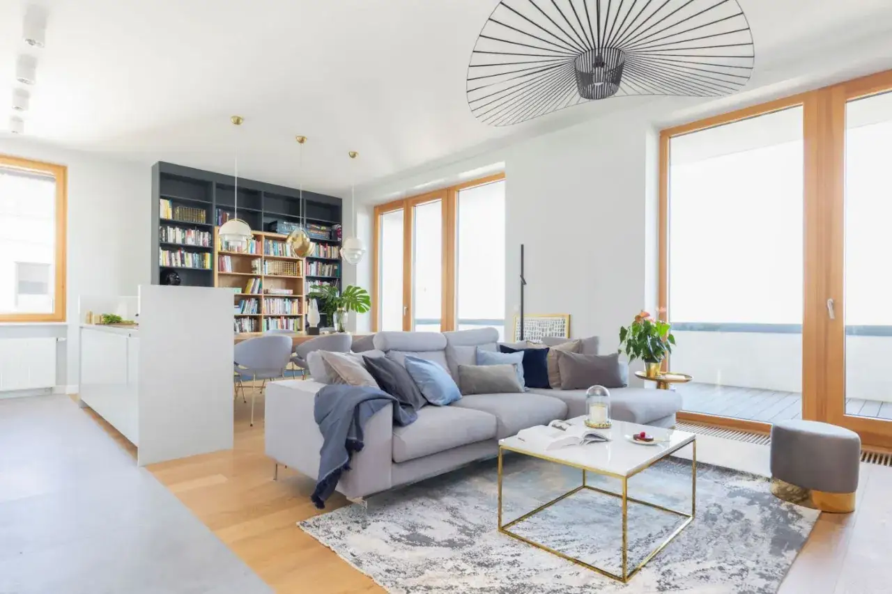 Nowoczesny salon z białymi ścianami. Szara sofa z poduszkami, marmurowy stolik kawowy, regał z książkami i designerska lampa. Jakie dodatki podkreślają styl?