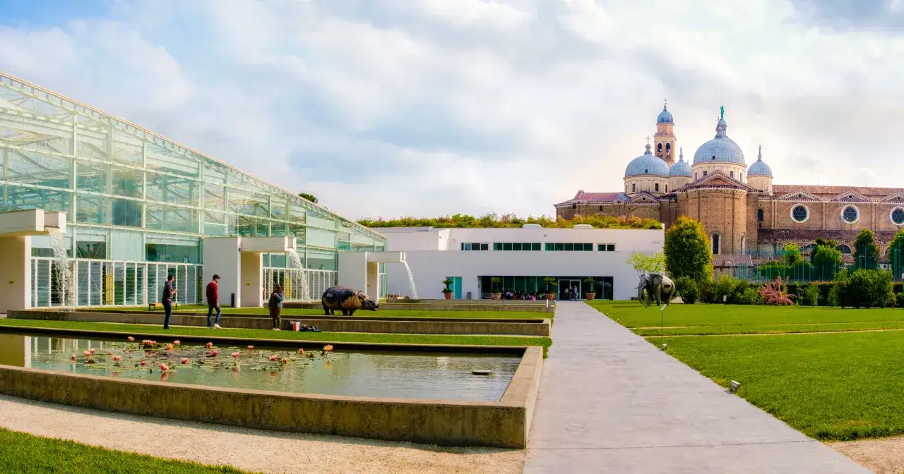 Ogród botaniczny w Padwie: nowoczesna szklarnia, stawy z liliami wodnymi, rzeźby zwierząt i zabytkowa bazylika w tle.