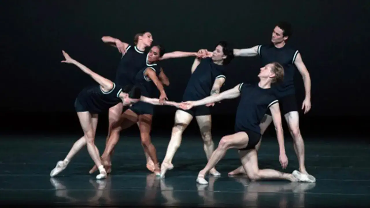 Sześcioro tancerzy w czarnych strojach tworzy dynamiczną choreografię, splatając ramiona w skomplikowanej sekwencji ruchów.