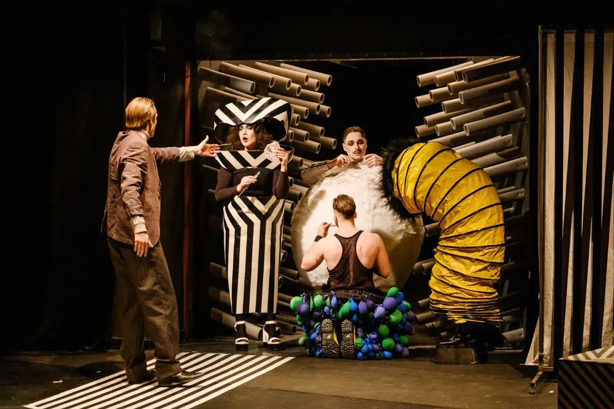 Scena teatralna z postaciami w surrealistycznych strojach, przypominająca dramaty mrożka alfabetycznie.