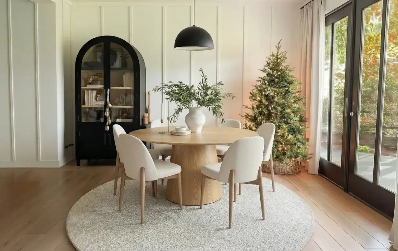 En vacker dukning med ett runt tr&auml;bord, beiga stolar, en julgran och ett svart sk&aring;p.
