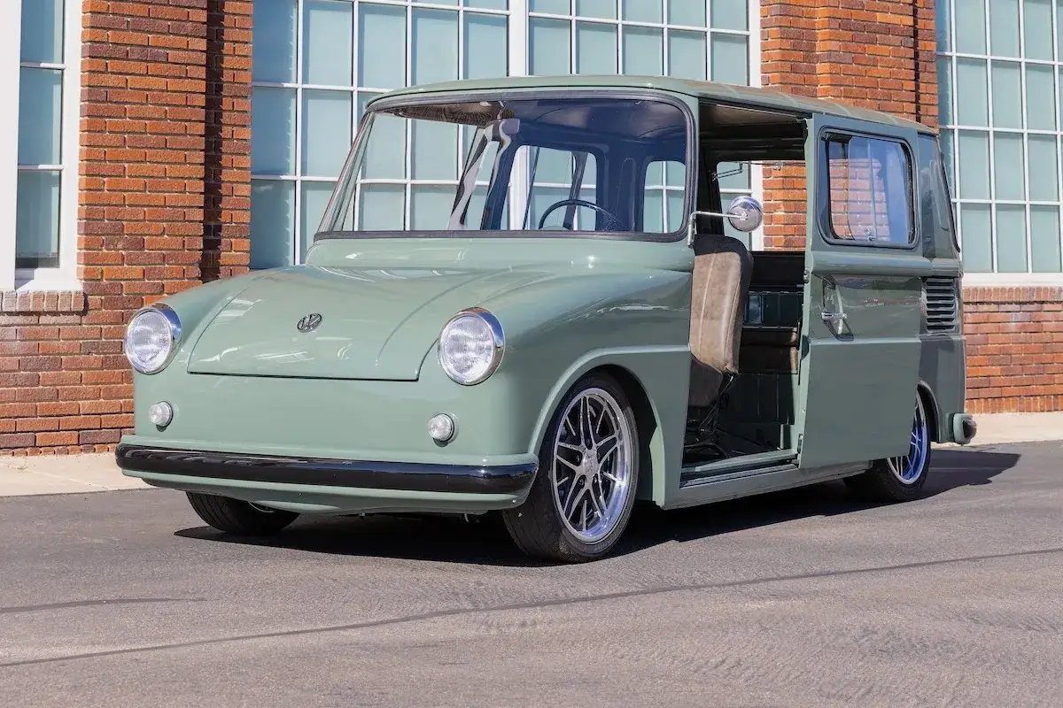 Ein grüner VW Fridolin mit geöffneter Tür steht vor einer Backsteinwand mit großen Fenstern.