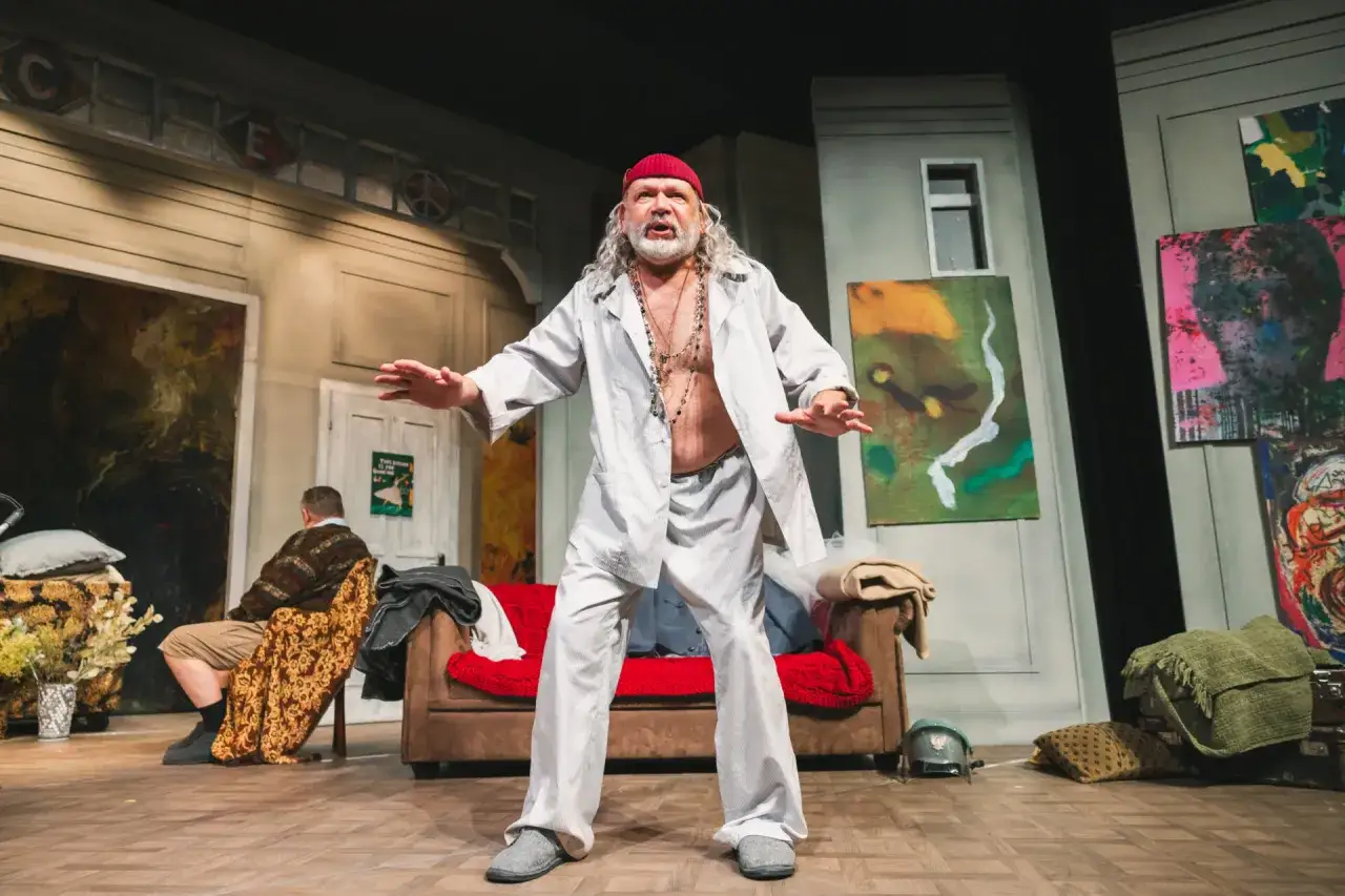 Aktor w czerwonej czapce i białej koszuli, z długimi włosami, gestykuluje na scenie. To scena z dramatu Mrożka.
