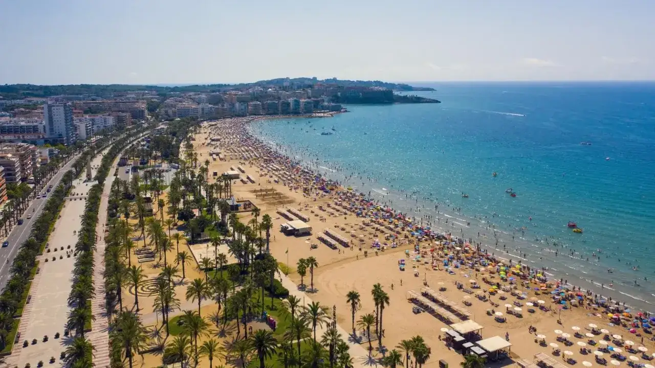 Tłoczna plaża na Costa Dorada, pełna ludzi i parasoli. W tle widać miasto i błękitne morze, idealne na wakacyjne atrakcje.