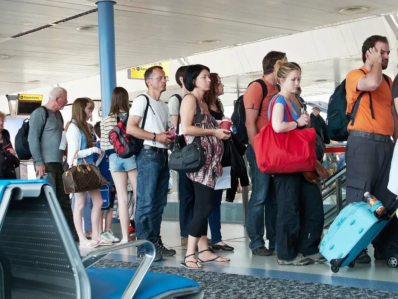 Ludzie w kolejce na lotnisku czekają na boarding. Wszyscy mają bagaże, niektórzy trzymają dokumenty.