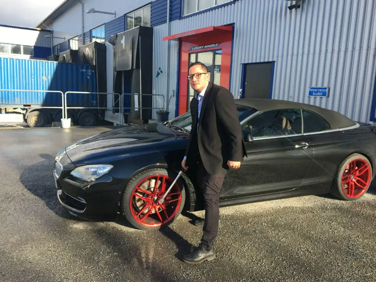 Man i kostym kontrollerar efterdra hjulbultar p&aring; en svart cabriolet med r&ouml;da f&auml;lgar.