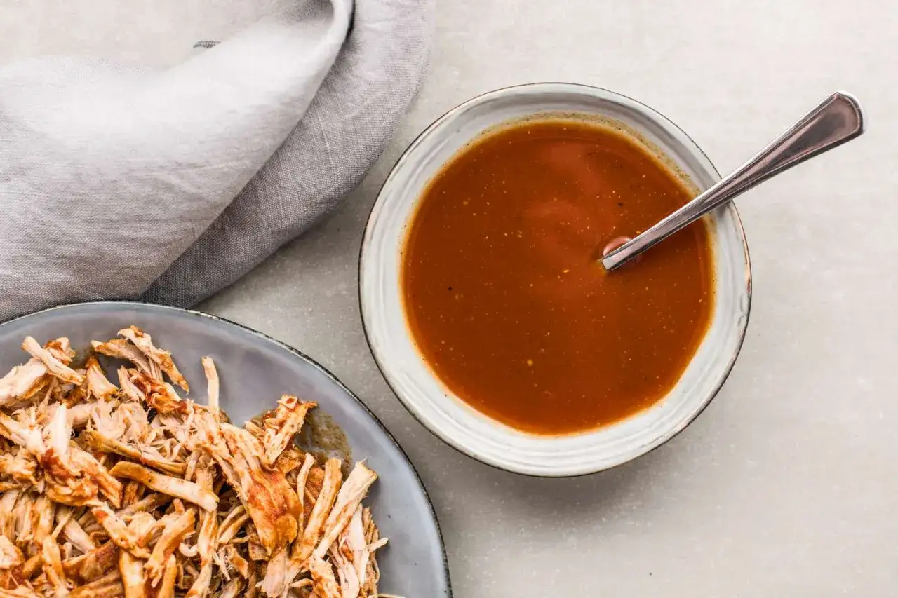 En sk&aring;l med mustig s&aring;s till pulled pork bredvid en tallrik med strimlat k&ouml;tt. En sked vilar i s&aring;sen.